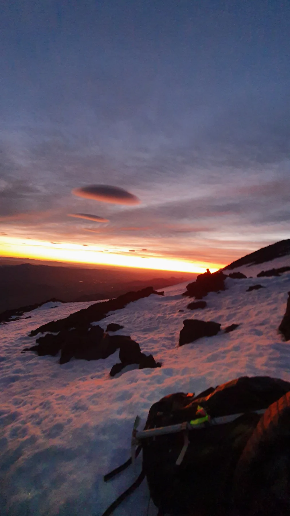 Sunrise from the upper slopes of Lanín, two lenticular clouds over the range, snow and volcanic rock in the foreground with an ice axe and pack