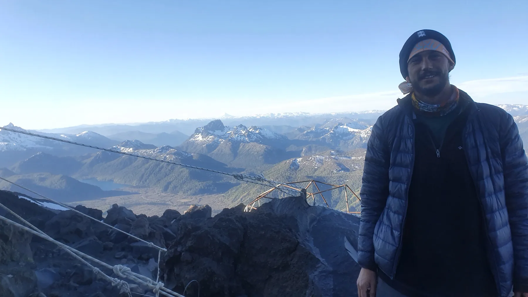 Standing at the CAJA refugio at 2,400 m on Lanín in evening light, steel guy wires and a hut frame in the foreground, snow-streaked peaks and a lake behind